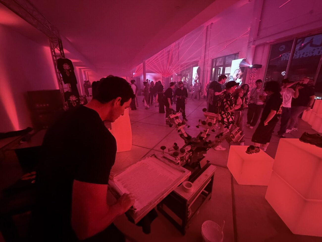 A printer flooding a screen with ink at a brightly lit event while a crowd lines up nearby