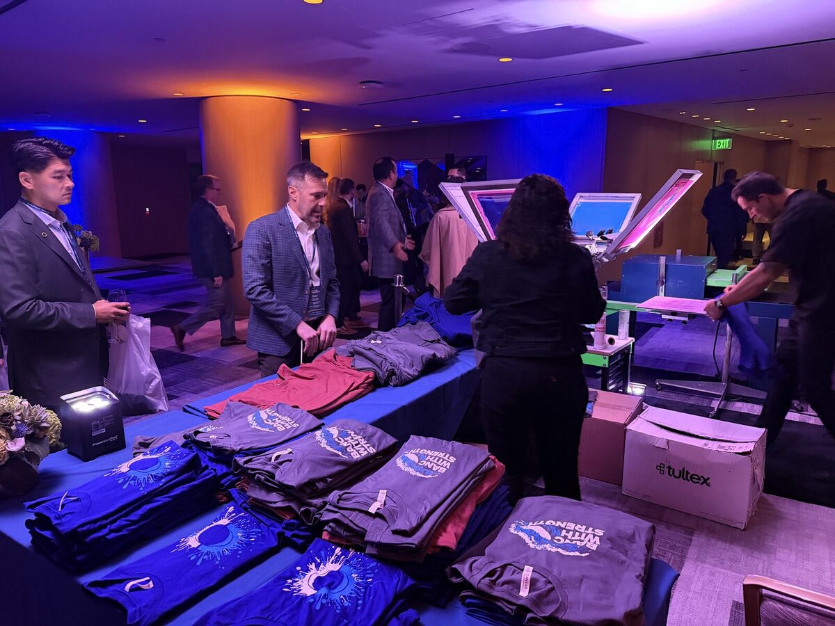 Folded freshly screen-printed event tees stacked on a table while a guest in a blazer looks on at a reception