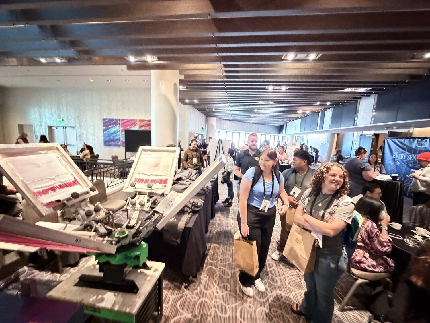 Merch Troop screen-printing station set up in a bright, window-lined event hall as smiling guests carry away freshly printed merch bags