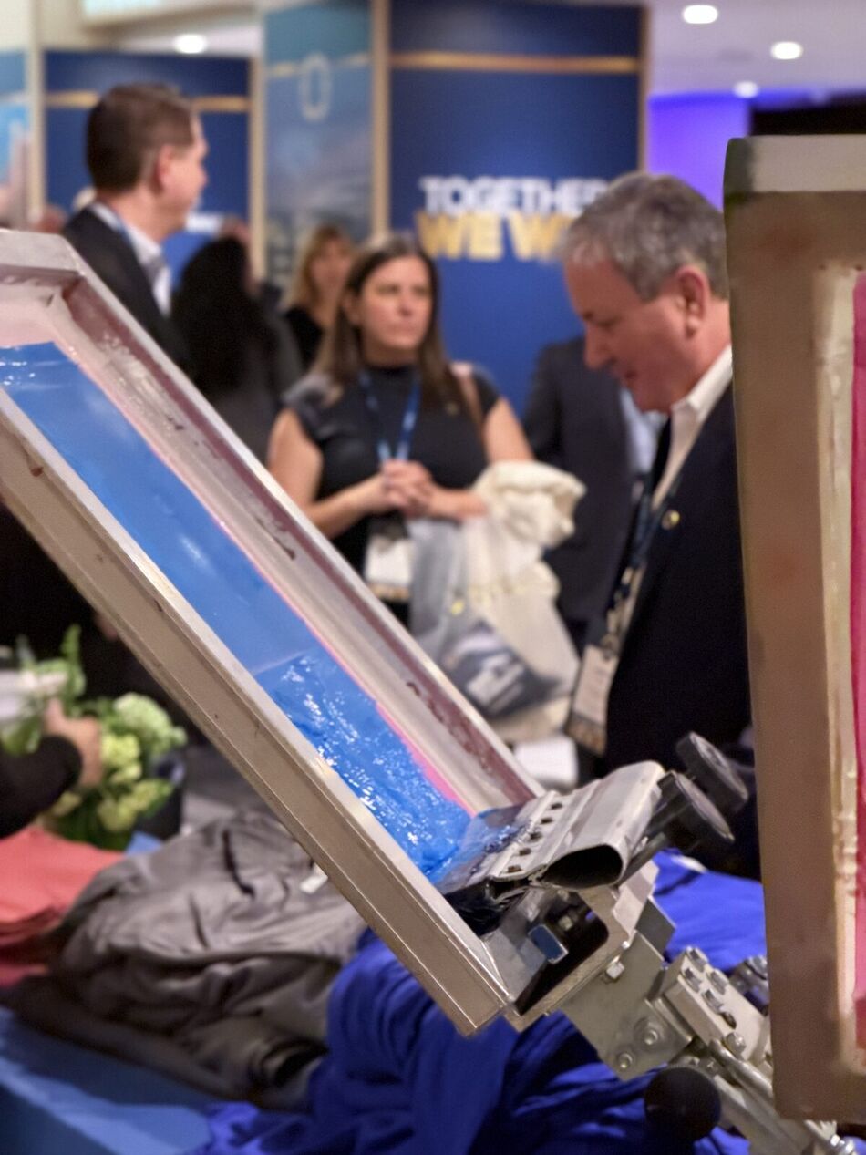 Close-up of blue ink spread across a screen-printing mesh frame with event guests gathered behind the booth